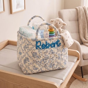 May include: A quilted fabric storage basket featuring a blue and white floral pattern with the name Robert embroidered in blue script on the front. The basket is filled with baby supplies including nappies, wipes, a tube of cream, a toy, and a plush lamb.