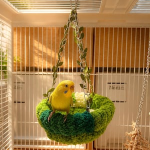 Puede incluir: Un periquito amarillo brillante posado dentro de un nido de pájaro redondo tejido a ganchillo. El nido presenta hilo texturizado verde y blanco, suspendido por cadenas de metal plateado adornadas con pequeñas hojas verdes artificiales.