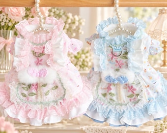 May include: Two pet dresses hanging on pearl-beaded hangers. One dress is pink with white lace ruffles, floral embroidery, and a white faux fur accent. The other dress is light blue with matching lace ruffles, floral embroidery, and a blue faux fur accent.