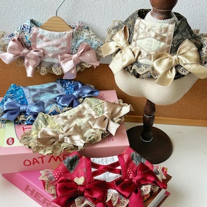 Puede incluir: Una colección de collares decorativos para mascotas en varios colores y diseños. Los collares presentan lazos, ribetes de encaje y patrones decorativos. Un collar se exhibe en una cabeza de maniquí de madera.