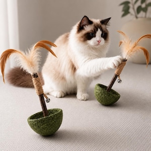 May include: Two cat toys, each featuring a green felted bowl base, a dark wooden stick wrapped in tan twine, a small silver bell, and light brown feathers at the top. A fluffy white and brown cat is reaching toward one of the toys.