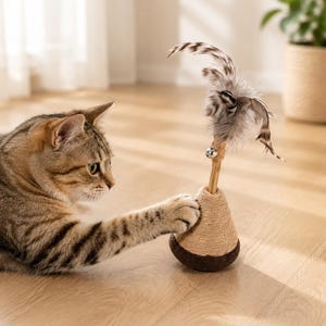 May include: A brown tabby cat reaches out with a paw to touch a cone-shaped cat toy. The toy features a tan sisal-wrapped base, a small silver bell, and a cluster of brown, white, and grey feathers attached to a central wooden stick.