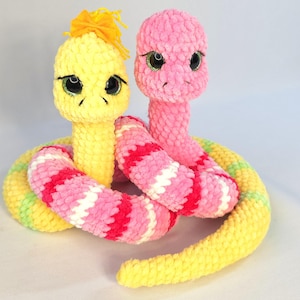 Puede incluir: Dos juguetes de serpiente de ganchillo. Uno es amarillo con un mechón de hilo amarillo en la cabeza, el otro es rosa. Sus cuerpos están entrelazados, con el cuerpo de la serpiente amarilla a rayas rosas, blancas y verdes. El cuerpo de la serpiente rosa está a rayas rosas y blancas.