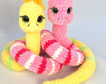 May include: Two crocheted snake toys. One is yellow with a yellow tuft of yarn on its head, the other is pink. Their bodies are intertwined, with the yellow snake's body striped with pink, white, and green. The pink snake's body is striped with pink and white.