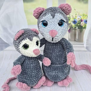 May include: Two handmade crochet possums. The possums are gray with pink ears, paws, and tails. They have blue eyes and pink noses. One possum is smaller than the other. The possums are standing on a white wooden surface.