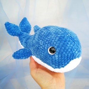 Pode incluir: Um brinquedo de pelúcia de baleia azul com detalhes brancos. A baleia tem uma aparência macia e texturizada, com uma barriga branca e olhos grandes e pretos. As barbatanas e a cauda também são azuis. O brinquedo é segurado contra um fundo azul claro.