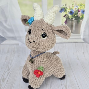 May include: A handmade, plush goat toy in tan with white horns and black hooves. It has a small bell necklace and a blue flower on its head, plus a red flower on its side. The toy is set against a window with flowers in the background.