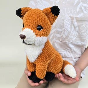 Può includere: Un peluche a forma di volpe all'uncinetto fatto a mano, arancione e bianco, con dettagli neri. La volpe ha petto, muso e punta della coda bianchi. La volpe è tenuta tra le mani di una persona. È un giocattolo morbido e coccoloso.
