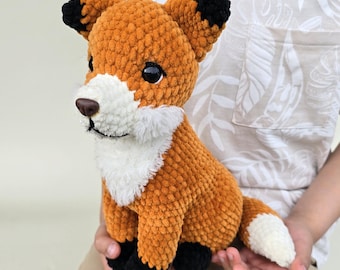 Könnte beinhalten: Ein handgefertigtes, gehäkeltes Fuchsplüschtier in Orange und Weiß mit schwarzen Akzenten. Der Fuchs hat eine weiße Brust, Schnauze und Schwanzspitze. Der Fuchs wird in den Händen einer Person gehalten. Das Kuscheltier ist weich.