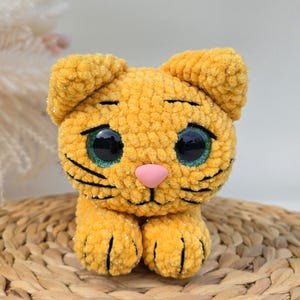 Może przedstawiać: Ręcznie robiony, pluszowy pomarańczowy kot z dużymi, błyszczącymi niebieskimi oczami i różowym nosem. Szydełkowy kot ma czarne wąsy i siedzi na plecionej macie. Uroczy, przytulny i dekoracyjny przedmiot.