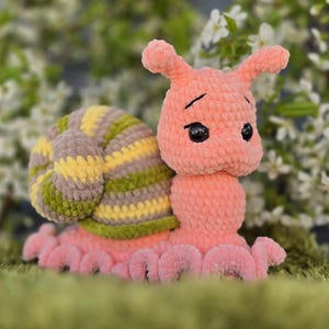 May include: A handmade crochet snail toy in shades of peach, yellow, green, and brown. The snail has a textured shell with stripes and a smiling face with black eyes. The snail is sitting on a green surface.