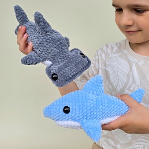 May include: Two plush shark toys. One is a light blue shark with a white belly, and the other is a gray hammerhead shark. Both have black button eyes and are made of a soft, textured material.
