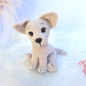 Könnte beinhalten: Ein hellbeige Plüschhundespielzeug mit großen, schlaffen Ohren und einer schwarzen Nase. Der Hund sitzt aufrecht mit den Pfoten nach vorne und hat große, dunkle Augen. Das Spielzeug besteht aus einem weichen, strukturierten Material.