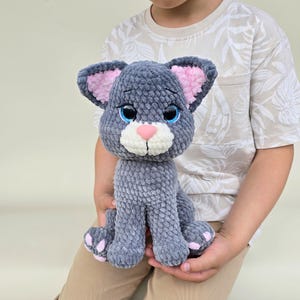 Puede incluir: Un peluche de gato gris tejido a ganchillo con orejas internas rosas, una nariz rosa y ojos azules. El animal de peluche hecho a mano tiene un hocico blanco y almohadillas rosas. El gato está sentado erguido y lo sostiene una persona.