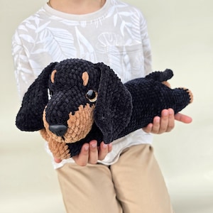 Puede incluir: Un perro salchicha de peluche negro y canela. El animal de peluche hecho a mano tiene un aspecto suave y texturizado con orejas caídas y un cuerpo largo. El perro es sostenido por una persona que lleva una camisa blanca y pantalones color caqui.