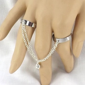May include: A silver ring set connected by a delicate chain. The set includes two rings, one on the index finger and one on the middle finger. The chain has a small charm at the end. The rings are displayed on a hand.