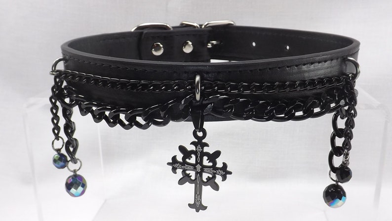 May include: Black leather choker with multiple black chains and a dangling silver cross pendant. The chains have iridescent blue beads at the ends.