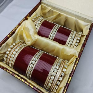 Puede incluir: Un juego de dos pulseras granates con detalles dorados y pedrería, presentado en una caja decorativa con forro de satén. Las pulseras son de color rojo oscuro y presentan detalles intrincados.