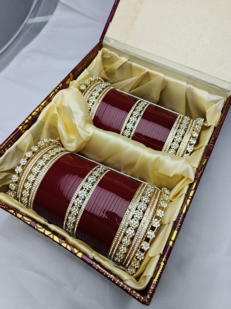 K&ouml;nnte beinhalten: Zwei weinrote Armreifen mit goldenen und Strass-Akzenten, pr&auml;sentiert in einer dekorativen Box mit Satinfutter. Die Armreifen haben eine tiefrote Farbe und weisen aufwendige Details auf.