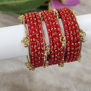 Può includere: Un set di braccialetti rossi con dettagli dorati e piccoli ciondoli pendenti. I braccialetti sono adornati con piccole pietre rosse quadrate. Sullo sfondo si vedono fiori viola.