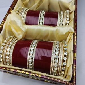 K&ouml;nnte beinhalten: Zwei weinrote Armreifen mit goldenen und Strass-Akzenten, pr&auml;sentiert in einer dekorativen Box mit goldenem Satinfutter. Die Armreifen liegen nebeneinander und zeigen ihr aufwendiges Design und ihre Handwerkskunst.