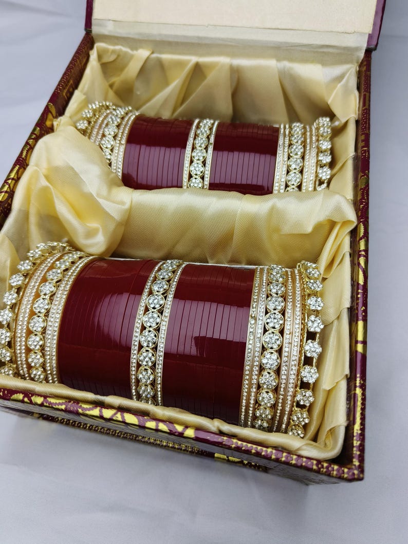 K&ouml;nnte beinhalten: Zwei weinrote Armreifen mit goldenen und Strass-Akzenten, pr&auml;sentiert in einer dekorativen Box mit goldenem Satinfutter. Die Armreifen liegen nebeneinander und zeigen ihr aufwendiges Design und ihre Handwerkskunst.