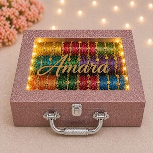 Könnte beinhalten: Eine rosa glitzernde Box mit durchsichtigem Deckel, gefüllt mit bunten Armreifen. Das Wort "Amara" ist in goldener Schrift dargestellt. Die Box hat einen silbernen Griff und Verschluss und wird von kleinen Lichtern beleuchtet.