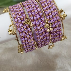 Può includere: Un set di braccialetti color lavanda con un motivo a cuore e dettagli dorati. Ogni braccialetto è adornato con piccoli ciondoli dorati. I gioielli sono esposti su una superficie bianca, con fiori viola sullo sfondo.