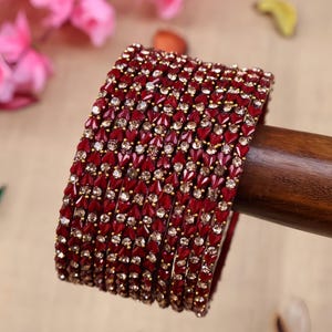 Può includere: Una pila di braccialetti rossi e dorati. I braccialetti sono adornati con piccole pietre rosse a forma di cuore e intervallati da accenti dorati. I gioielli sono esposti su un tassello di legno, con uno sfondo sfocato.