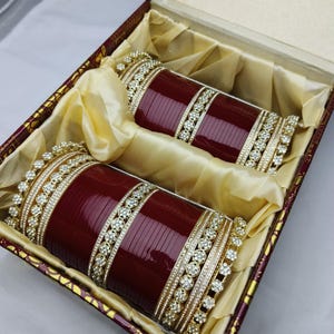 K&ouml;nnte beinhalten: Zwei weinrote Armreifen mit goldenen und Strass-Akzenten, pr&auml;sentiert in einer dekorativen Box. Die Armreifen liegen auf goldenem Stoff, der ihr detailliertes Design hervorhebt. Die Box hat eine weinrote Au&szlig;enseite mit goldenen Verzierungen.