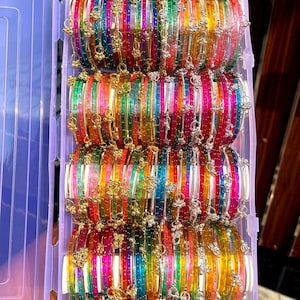 Puede incluir: Una colección de pulseras coloridas en un recipiente de plástico transparente. Las pulseras son una mezcla de colores vibrantes, incluyendo rojo, verde, amarillo y azul, con detalles decorativos. Las pulseras están apiladas ordenadamente, mostrando su intrincado diseño.