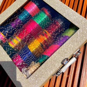 Puede incluir: Una caja decorativa con tapa transparente llena de pulseras de cristal de colores. Las pulseras son en tonos rosa, verde, azul, amarillo y morado. La caja tiene un marco dorado brillante y un cierre plateado.