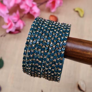 Könnte beinhalten: Ein Stapel türkisfarbener Armreifen mit goldenen und klaren Akzenten. Die Armreifen sind mit kleinen, funkelnden Verzierungen versehen. Der Schmuck wird auf einem Holzstab präsentiert, mit rosa Blumen im Hintergrund.
