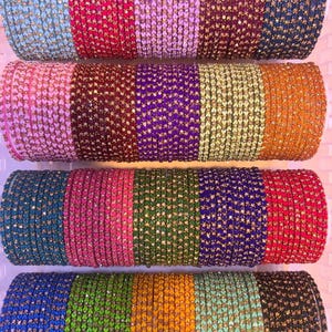 Op de afbeelding: Rijen kleurrijke armbanden in verschillende tinten, waaronder blauw, roze, paars en goud. Elke armband is versierd met kleine, sprankelende versieringen. De armbanden zijn op elkaar gestapeld en tonen een verscheidenheid aan kleuren en ontwerpen.