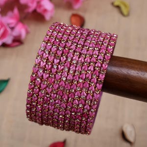 May include: A stack of pink bangles with heart-shaped embellishments and gold accents. The bangles are displayed on a wooden dowel, with pink flowers in the background. The bangles are approximately 0.25 inches wide.