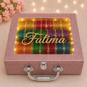 Könnte beinhalten: Eine rosa glitzernde Schmuckschatulle mit durchsichtigem Deckel, gefüllt mit bunten Armreifen. Die Box hat einen silbernen Griff und Verschluss. Der Name "Fatima" ist in goldener Schrift im Inneren zu sehen, beleuchtet von kleinen Lichtern.