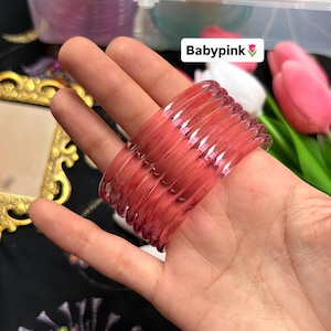 Könnte beinhalten: Ein Stapel durchscheinender, babyrosa Armreifen. Die Armreifen werden in einer Hand gehalten, mit dem Text "Babypink" in einer weißen Box mit einer rosa Blume. Im Hintergrund sind weitere Armreifen in verschiedenen Farben zu sehen.