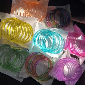 Op de afbeelding: Een verzameling kleurrijke, transparante armbanden in diverse tinten, waaronder oranje, geel, groen, blauw, roze en paars. Elke set is verpakt in een doorzichtige plastic zak.