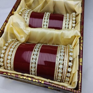 K&ouml;nnte beinhalten: Zwei Sets weinroter Armreifen mit goldenen und Strass-Akzenten, pr&auml;sentiert in einer dekorativen Box. Die Armreifen liegen auf goldenem Stoff, der ihr aufwendiges Design hervorhebt.