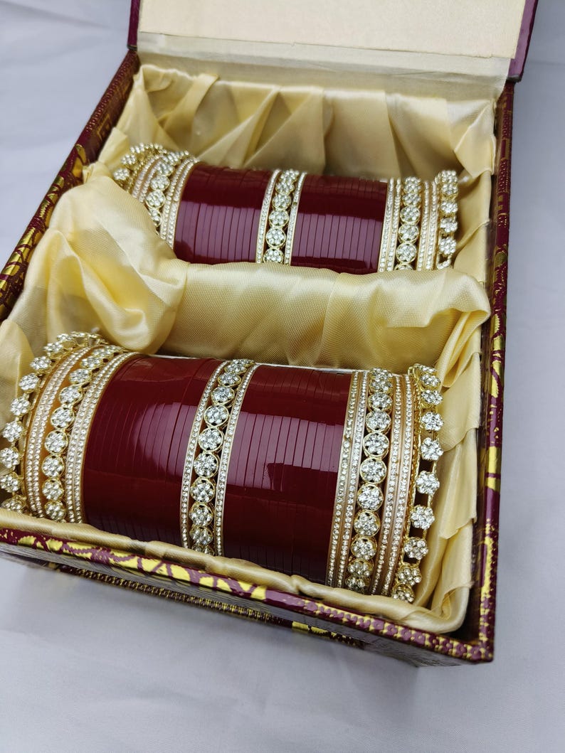 K&ouml;nnte beinhalten: Zwei Sets weinroter Armreifen mit goldenen und Strass-Akzenten, pr&auml;sentiert in einer dekorativen Box. Die Armreifen liegen auf goldenem Stoff, der ihr aufwendiges Design hervorhebt.