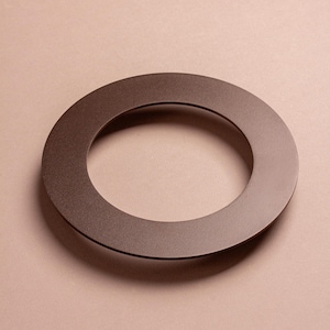 Könnte beinhalten: Ein braunes, kreisförmiges Metallobjekt mit einem Loch in der Mitte. Das Objekt hat eine matte Oberfläche und ist vor einem neutralen Hintergrund platziert. Die Form ist ein perfekter Ring, was auf einen dekorativen oder funktionalen Artikel hindeutet.