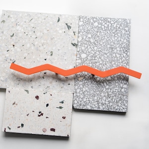 Può includere: Una maniglia arancione a forma di onda è posizionata su quattro piastrelle rettangolari in terrazzo nei toni del grigio e del bianco. Le piastrelle presentano un motivo maculato con vari aggregati colorati.