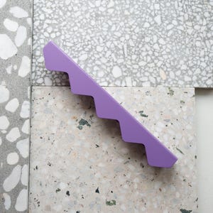 Pode incluir: Um objeto geométrico lavanda com uma borda escalonada. Está posicionado diagonalmente sobre uma superfície composta por ladrilhos de terrazzo em tons de cinza e branco, com um padrão salpicado.