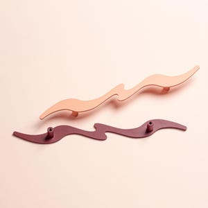 Könnte beinhalten: Zwei dekorative Schrankgriffe in wellenförmigem Design. Einer ist hellpfirsichfarben, der andere tief burgunderrot. Die Griffe haben eine moderne, minimalistische Ästhetik und sind auf hellrosa Hintergrund montiert.