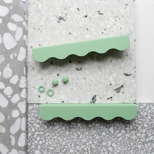 Könnte beinhalten: Ein Set aus mintgrünen Schrankgriffen und -knöpfen. Zwei wellenförmige Griffe sind oberhalb und unterhalb eines Sets aus drei runden Knöpfen und zwei kreisförmigen Unterlegscheiben angeordnet. Die Hardware wird auf einer gesprenkelten grau-weißen Oberfläche präsentiert.