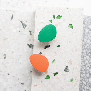 Pode incluir: Dois puxadores de armário ovais, um verde brilhante e outro laranja, repousam sobre uma superfície branca e salpicada. Os puxadores são lisos e sólidos, oferecendo um toque de cor contra o fundo neutro. Os puxadores são provavelmente para uma gaveta ou armário.