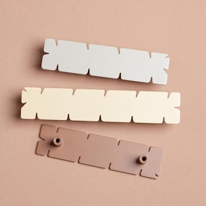 Può includere: Tre pomelli decorativi in metallo bianco, crema e bronzo. Ogni pomello ha una forma rettangolare con sezioni ritagliate. Il pomello in bronzo ha due fori per viti neri.