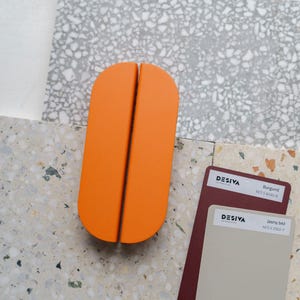 Puede incluir: Un objeto naranja en forma de píldora, dividido por la mitad, descansa sobre una superficie moteada. Se ven dos muestras de color de la marca Desiva, una burdeos y otra beige claro. El fondo presenta una mezcla de superficies moteadas y estampadas.