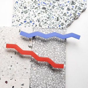 Op de afbeelding: Twee golvende, zigzagvormige haarclips. De ene is felrood, de andere is felblauw. De clips zijn geplaatst tegen een achtergrond van gespikkelde, getextureerde tegels in tinten grijs, wit en blauw.