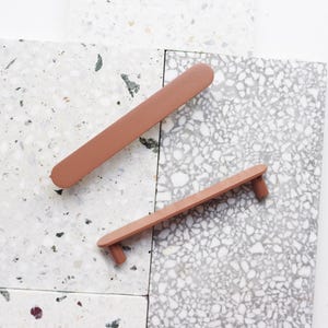 Puede incluir: Dos tiradores de armario de color terracota sobre una superficie moteada gris claro y blanca. Uno es una barra larga y rectangular, y el otro tiene una barra más corta con dos postes pequeños y redondeados. Herrajes para la decoración del hogar.